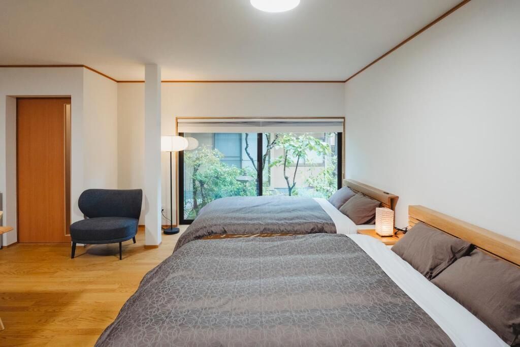 A bedroom with two beds, a chair, a bedside table, and a window with a view of trees.