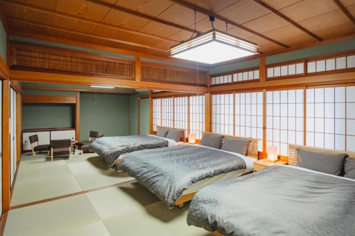 A traditional Japanese-style room with tatami flooring, a low ceiling, and sliding doors. There are three beds with grey bedding and a small table with chairs in the corner.