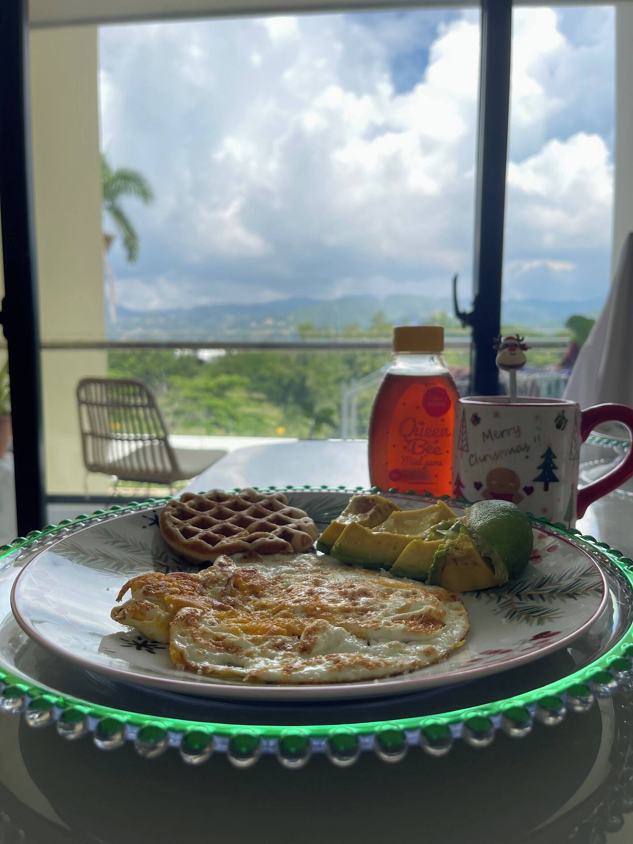 A breakfast plate with waffles, scrambled eggs, and avocado on a table with a green rim, overlooking a scenic view.