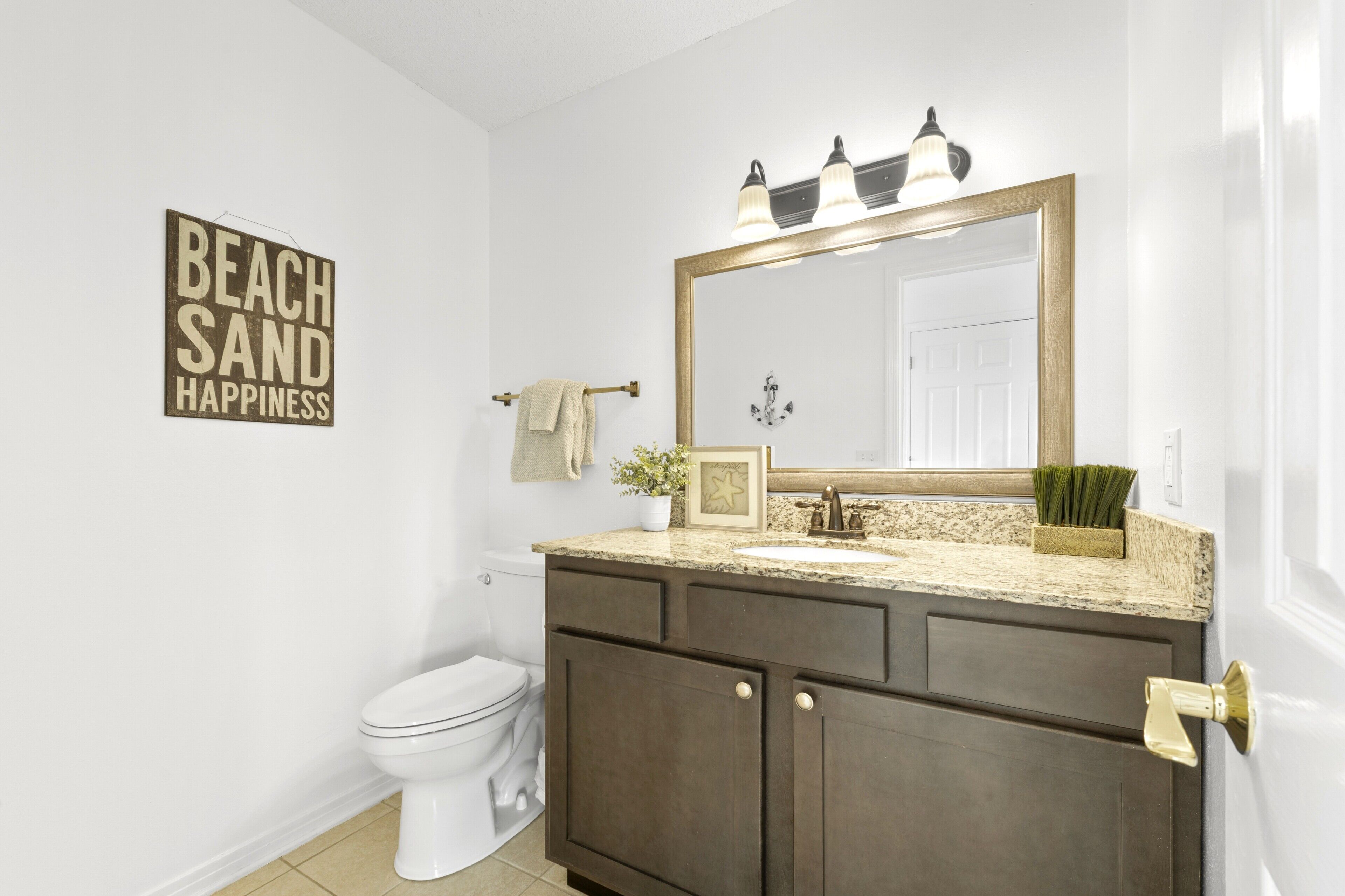 Baño con inodoro blanco, tocador de madera oscura, espejo grande con iluminación y un cartel que dice 'Beach Sand Happiness'.
