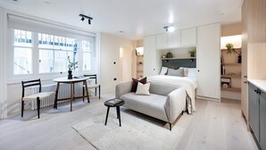 Un dormitorio moderno y bien iluminado con una cama grande, un sofá, una zona de comedor con mesa y sillas, y vistas al exterior a través de amplios ventanales.