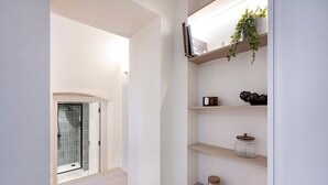 Un pasillo con una puerta de cristal que da al baño, estantes con adornos y una planta a la derecha.