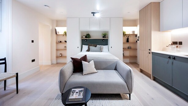 Un dormitorio moderno con una cama, un sofá gris y una mesita de noche con un libro. Se observa una zona de cocina con armarios y una encimera.