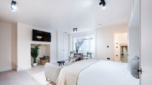Un dormitorio moderno con una cama grande, un televisor en la pared, una zona de comedor con mesa y sillas, y vistas a la ciudad a través de amplios ventanales.