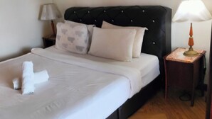 Cama bem arrumada com lençóis brancos e toalha dobrada, cabeceira estofada preta, mesa de cabeceira pequena com abajur e um quadro na parede.