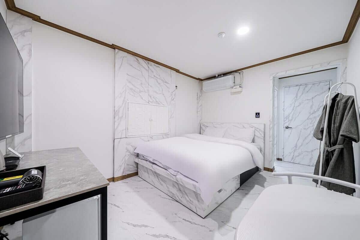 A modern bedroom with a marble-patterned wall, a bed, a bedside table with a kettle, and a robe hanging on a rack.