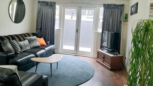 Ein Wohnzimmer mit schwarzem Ledersofa, einem Couchtisch, einem Flachbildfernseher auf einer Holzkommode und einer großen Zimmerpflanze.