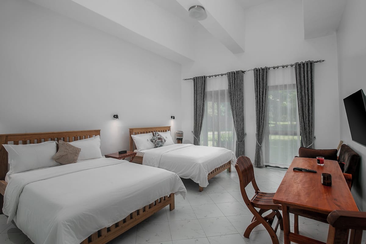 A hotel room with two single beds, a wooden table, a TV, and a large window with curtains.