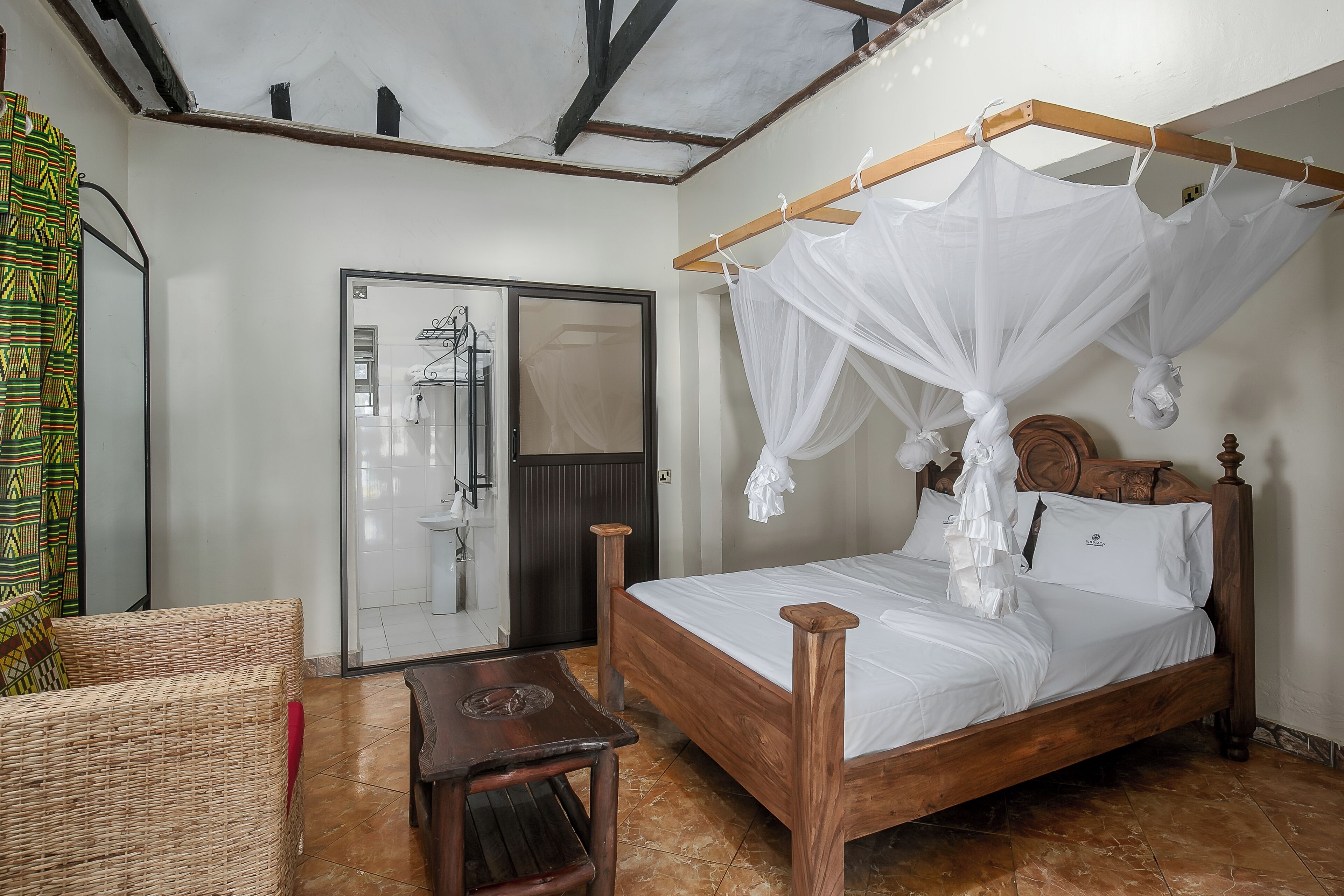 Un dormitorio con una cama de madera, una mesita de noche de madera, una silla de mimbre, un ventanal de cristal y un dosel blanco sobre la cama.