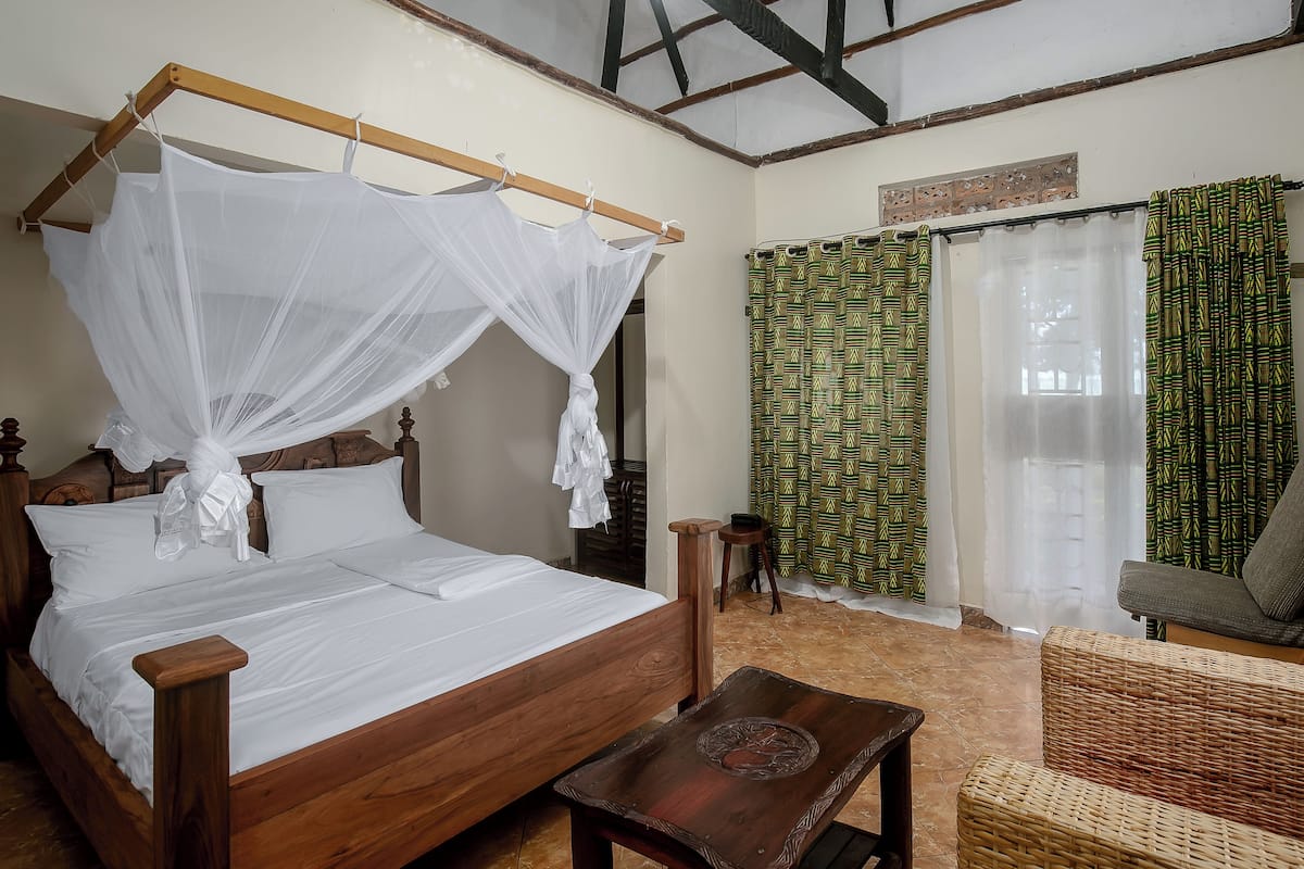 A wooden bed with a canopy, a small wooden table, and a wicker sofa in a room with patterned curtains and a tiled floor.