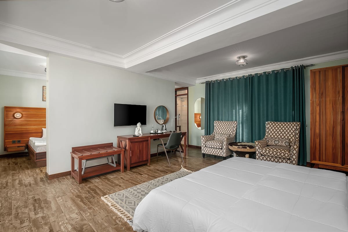 A hotel room with a large bed, wooden desk, two armchairs, a TV, and a mirror.