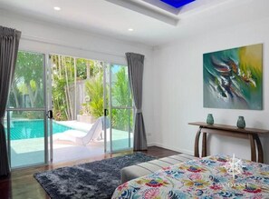 Ein Schlafzimmer mit einem großen Bett, einem Gemälde, einem Schreibtisch und Blick auf einen Pool und Grünflächen.