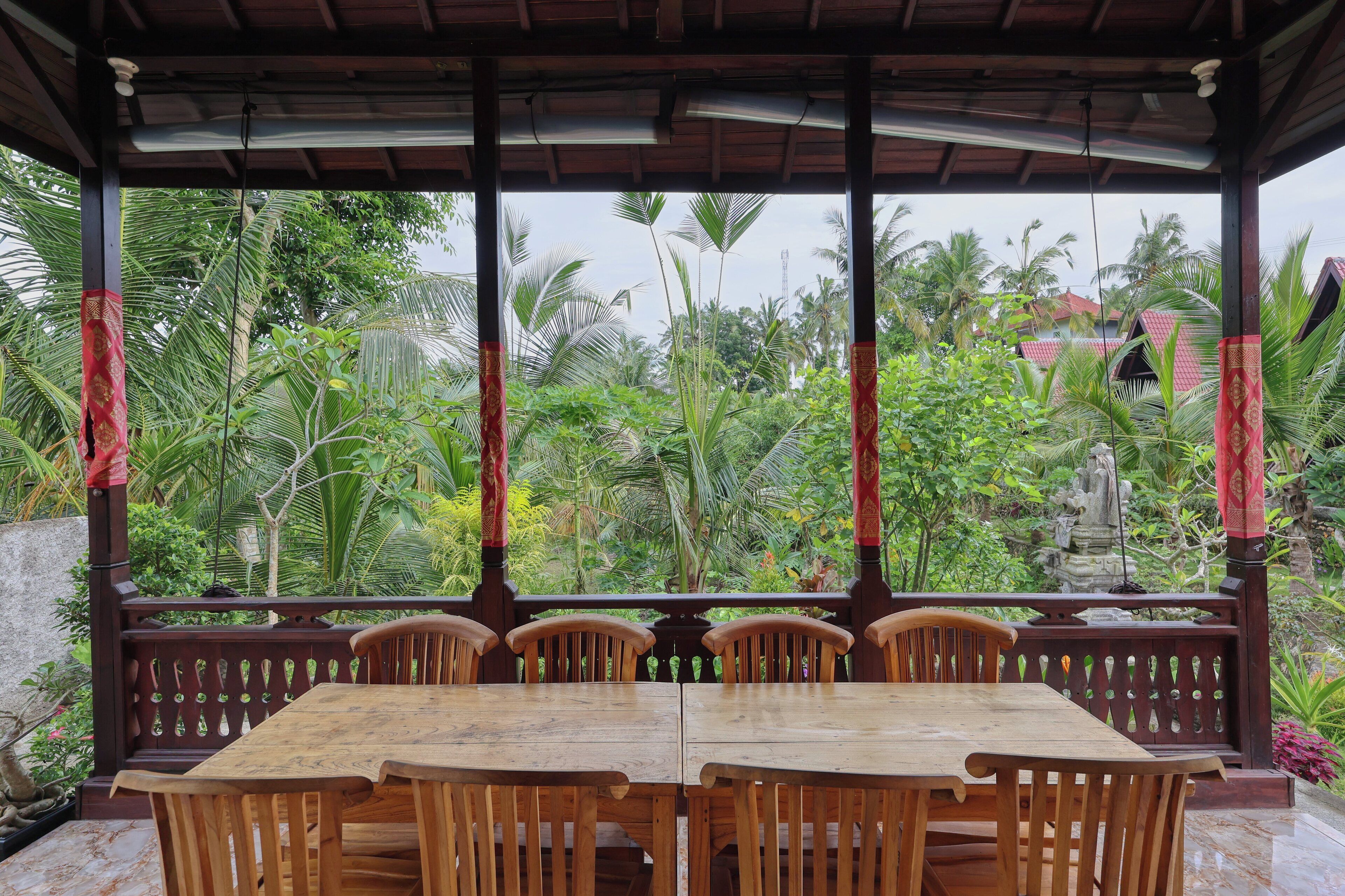 A covered outdoor area with a wooden table and chairs, surrounded by lush greenery and a stone pathway.
