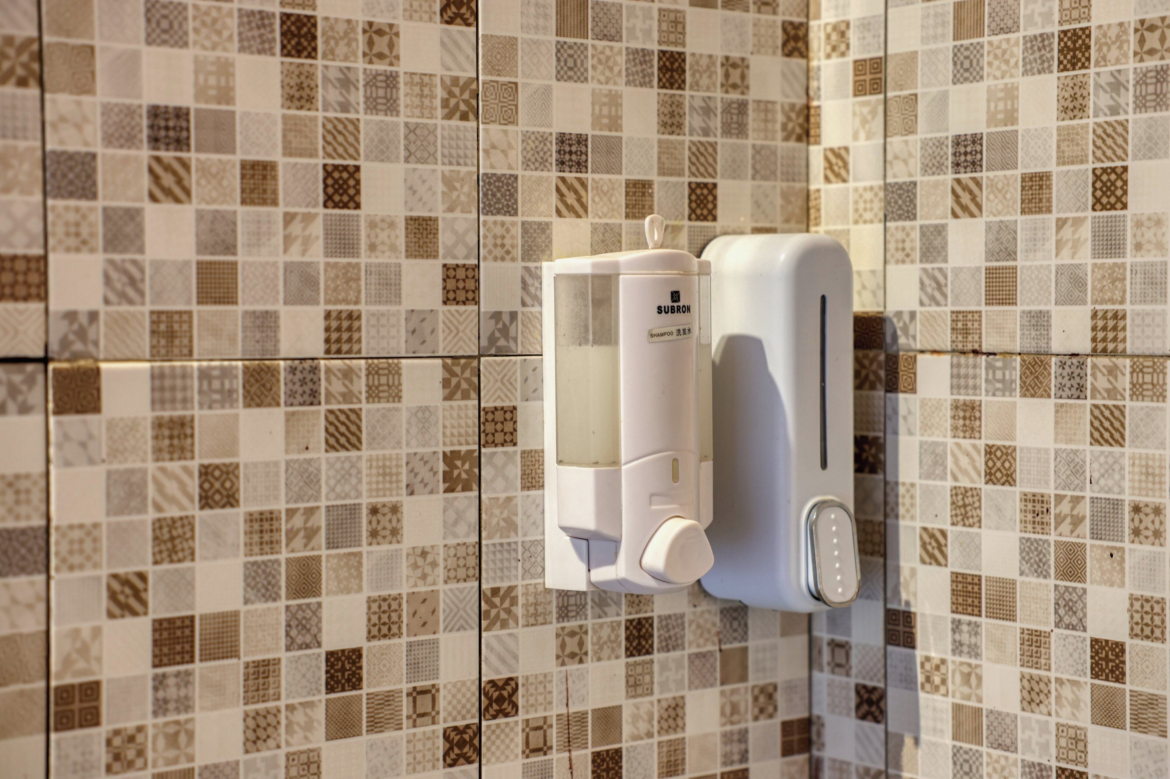 Dispensador de jabón montado en la pared con botella transparente y bomba, frente a una pared de azulejos con diversos diseños geométricos.
