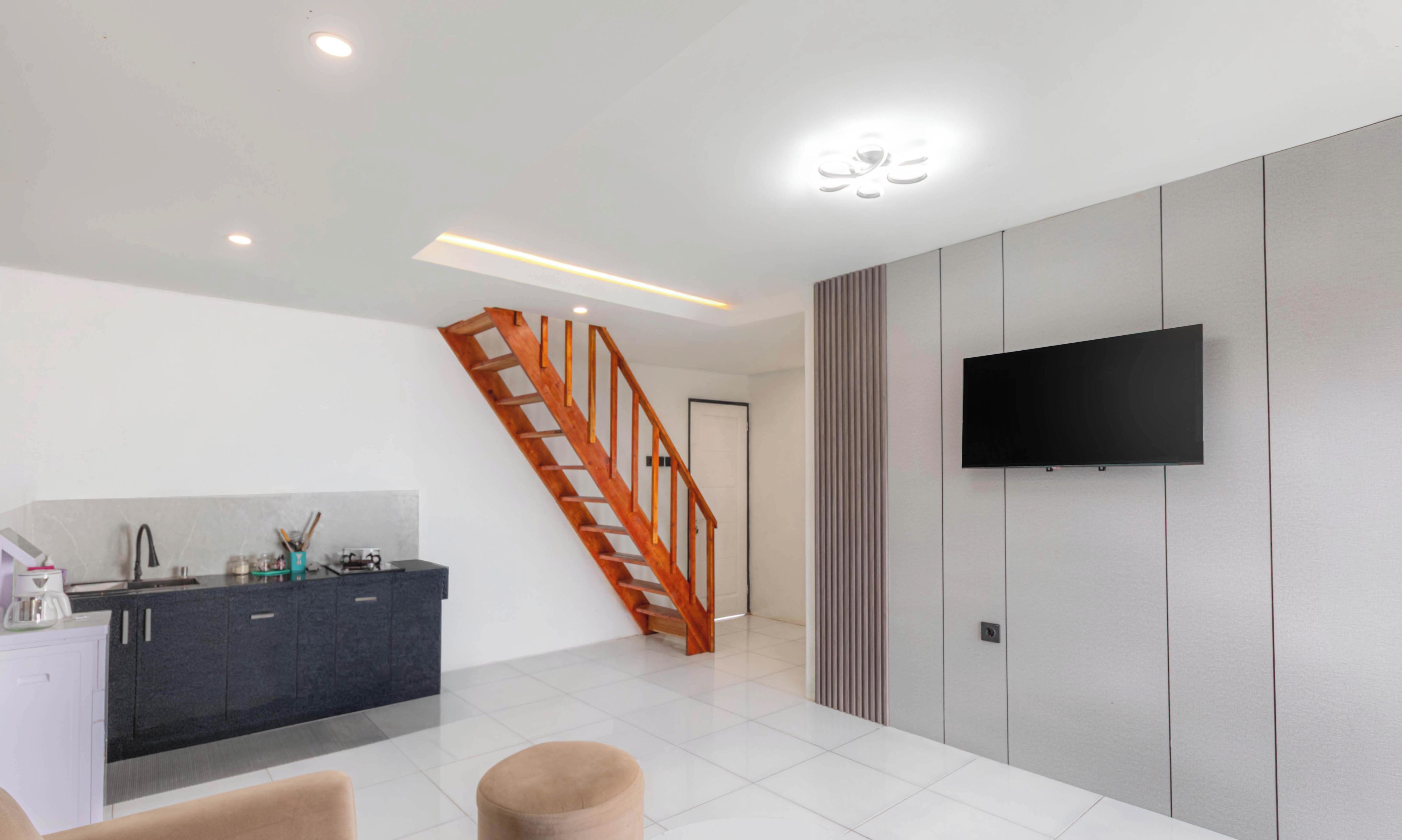 A modern living room with a staircase, a flat-screen TV mounted on the wall, and a kitchen area with a dark countertop.