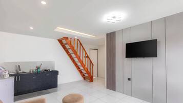 A modern living room with a staircase, a flat-screen TV mounted on the wall, and a kitchen area with a dark countertop.