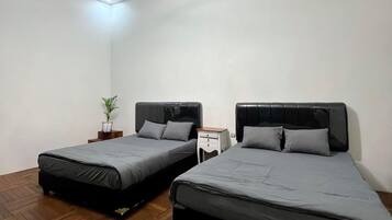 A room with two beds, a wooden herringbone floor, a ceiling light fixture, and a small bedside table with a plant.