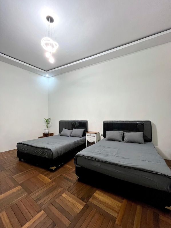Ein Zimmer mit zwei Betten, Holzboden im Fischgrätmuster, einer Deckenleuchte und einem kleinen Nachttisch mit einer Pflanze.