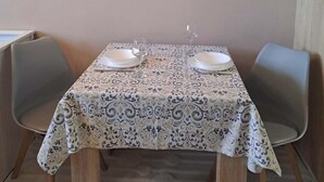 Un comedor con una mesa dispuesta para dos personas, dos sillas y una estantería en la pared con objetos decorativos.