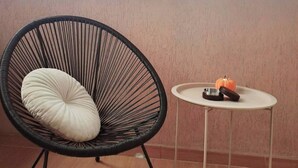 Una habitación con una silla de estructura negra y cojín blanco, una pequeña mesa con una calabaza y un control remoto, y una ventana con vista al cielo.