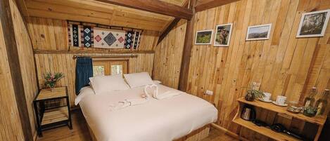 A cozy wooden room with a bed, a small table with a vase, and a shelf with items.