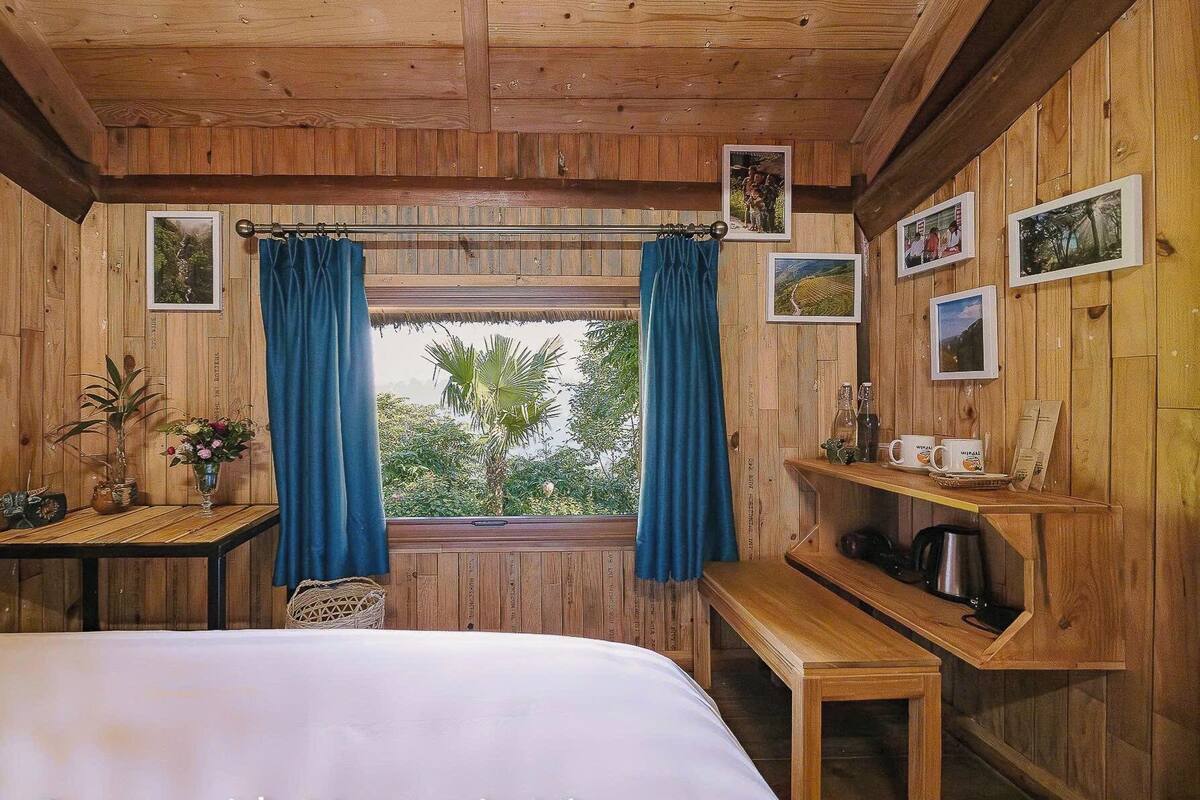 Una camera in una baita di legno con un letto, un tavolo con un vaso di fiori, una finestra con tende e uno scaffale con tazze e un bollitore.