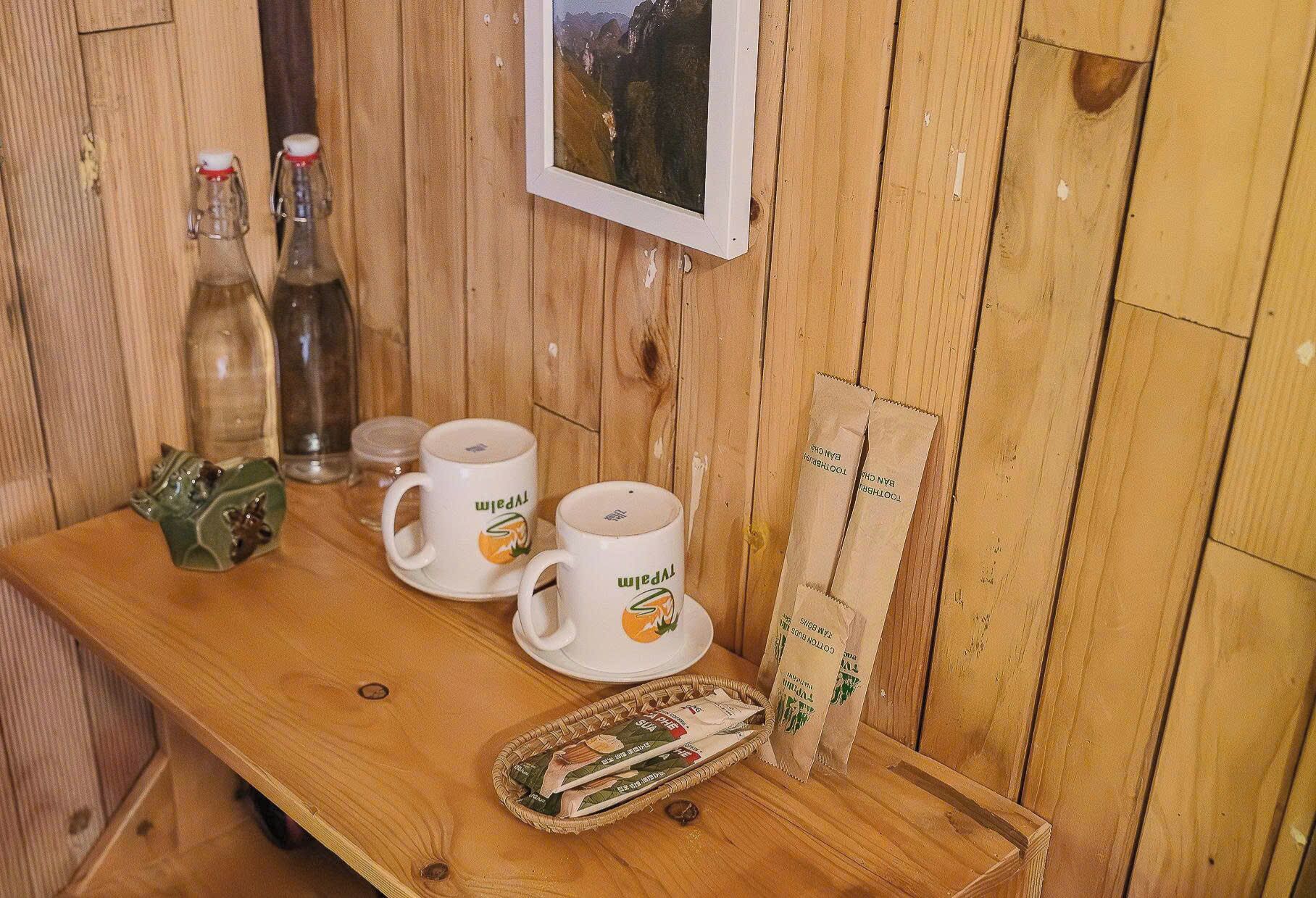 Une table en bois sur laquelle reposent deux tasses à café blanches, une figurine de grenouille en céramique, une bouteille en verre et un tableau encadré accroché au mur.