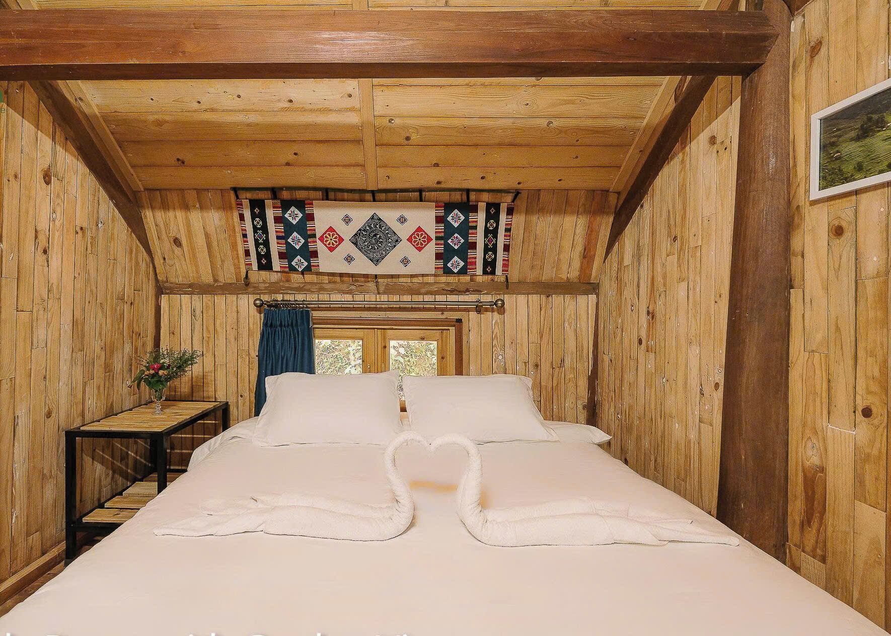 Ein Zimmer in einer Holzhütte mit einem Bett, einem kleinen Tisch mit einer Vase und einem Fenster mit Vorhängen.