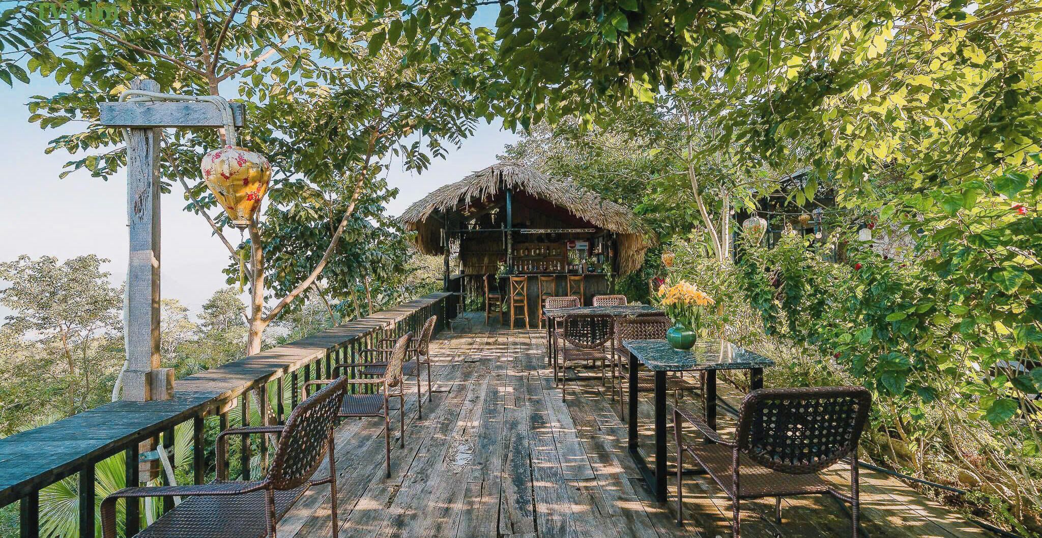 A wooden deck with wicker chairs and tables, leading to a thatched-roof structure with a bar area.