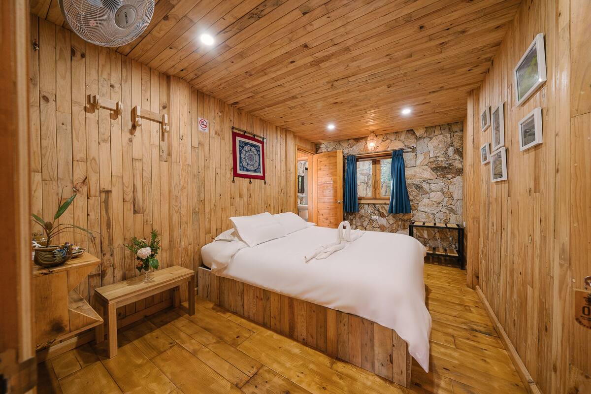 Una stanza con rivestimento in legno, un letto, una panca, un ventilatore e una parete in pietra.