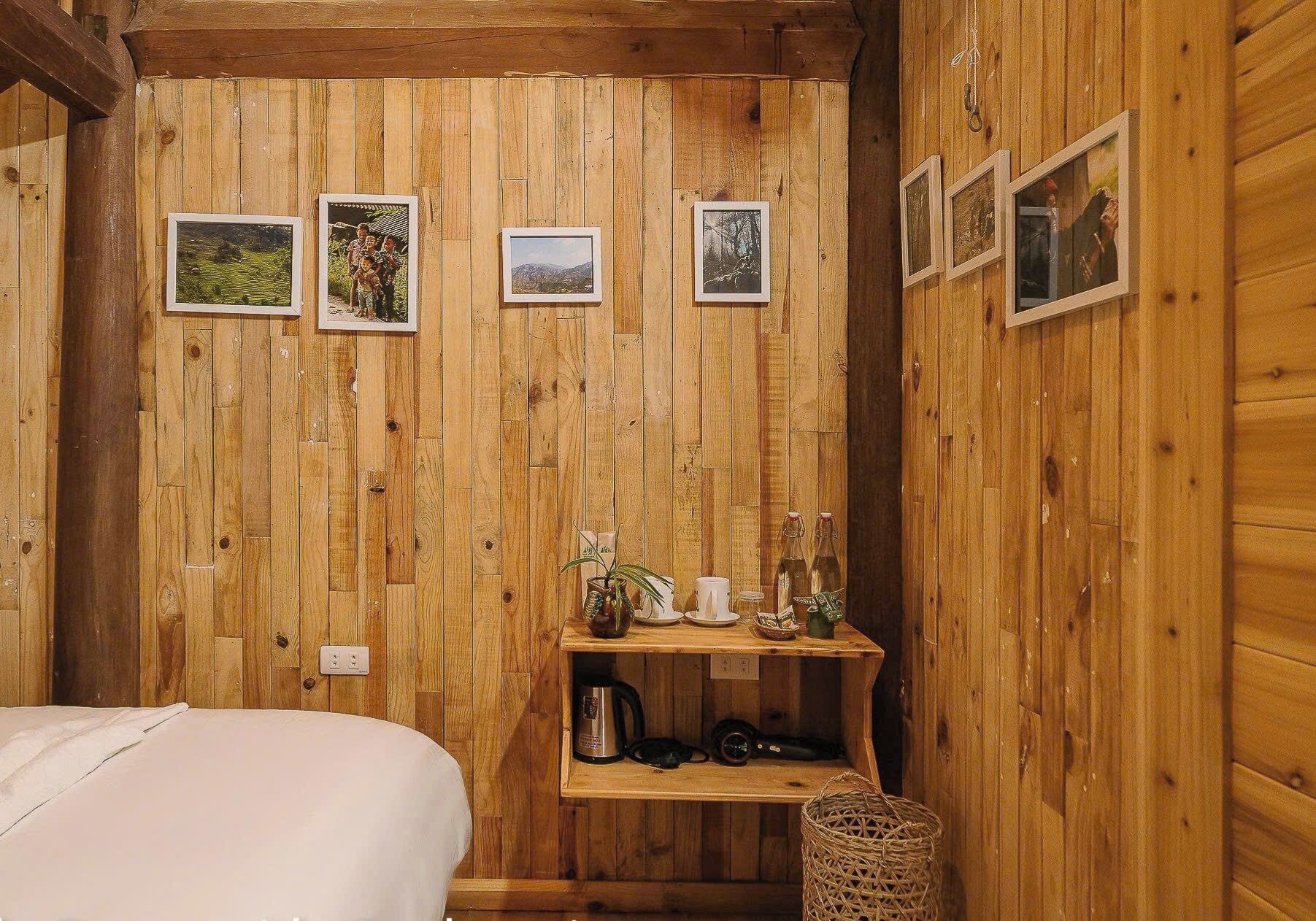 A wooden cabin room with a bed, a shelf with items, and three framed pictures on the wall.