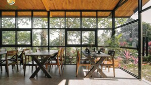A covered outdoor dining area with wooden tables and chairs, large windows, and a view of greenery. - TV Palm Ecolodge Ha Giang (Ha Giang 1)