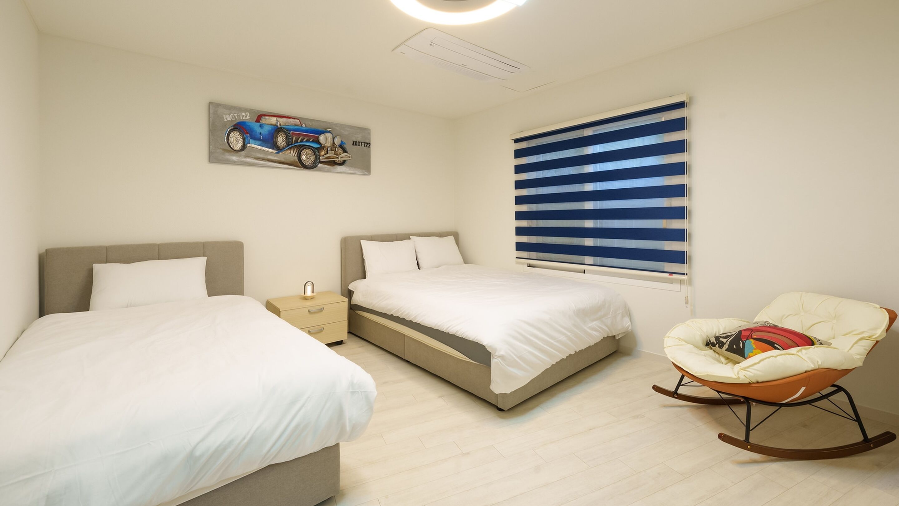 A bedroom with two beds, a rocking chair, a bedside table, a window with blinds, and a painting on the wall.