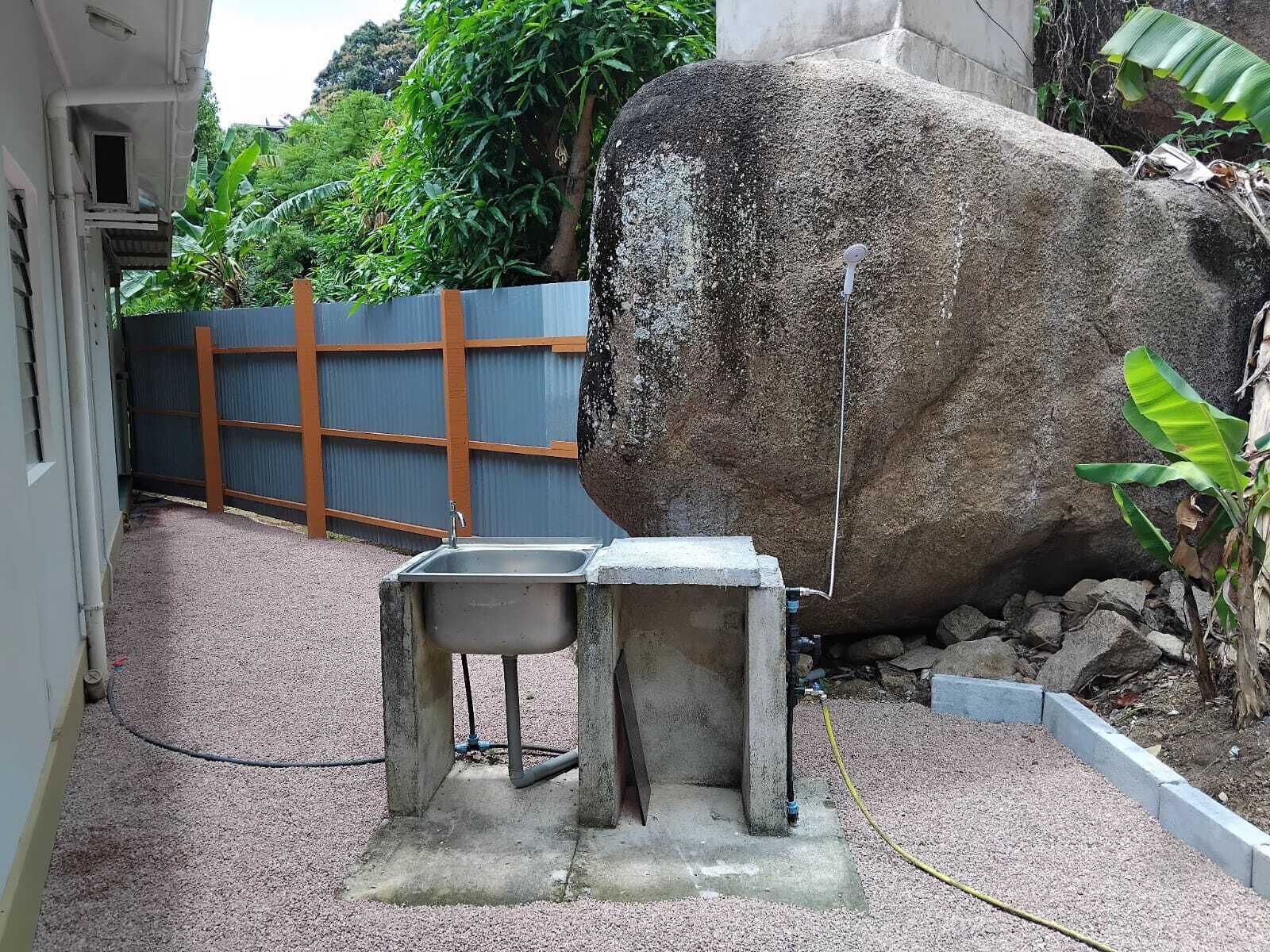 Un évier en béton avec un tuyau d’arrosage, un gros rocher et une clôture en métal dans un coin du jardin.
