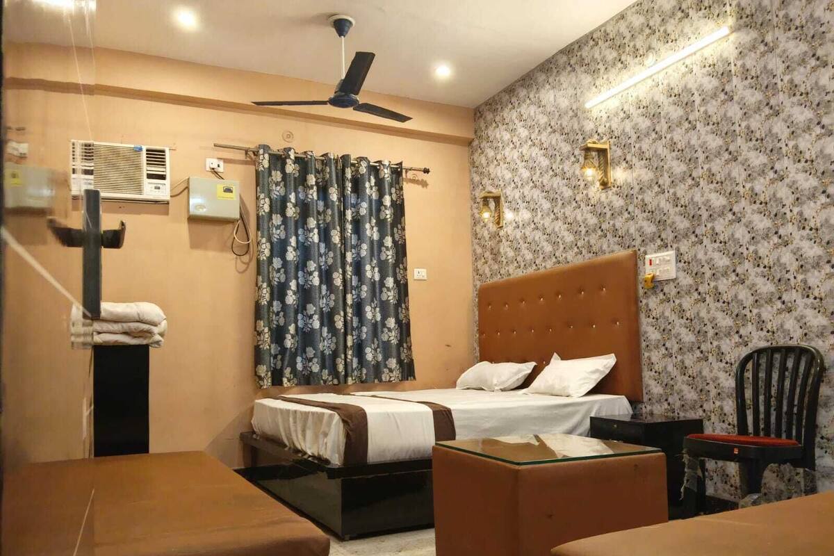 Habitación de hotel con cama, ventilador de techo, ventana con cortinas florales, una mesita y una silla.