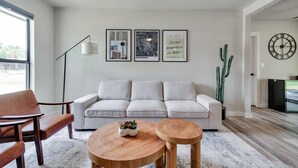 Una sala moderna con un sofá de color claro, dos mesitas de madera, una lámpara de pie y una planta de cactus.