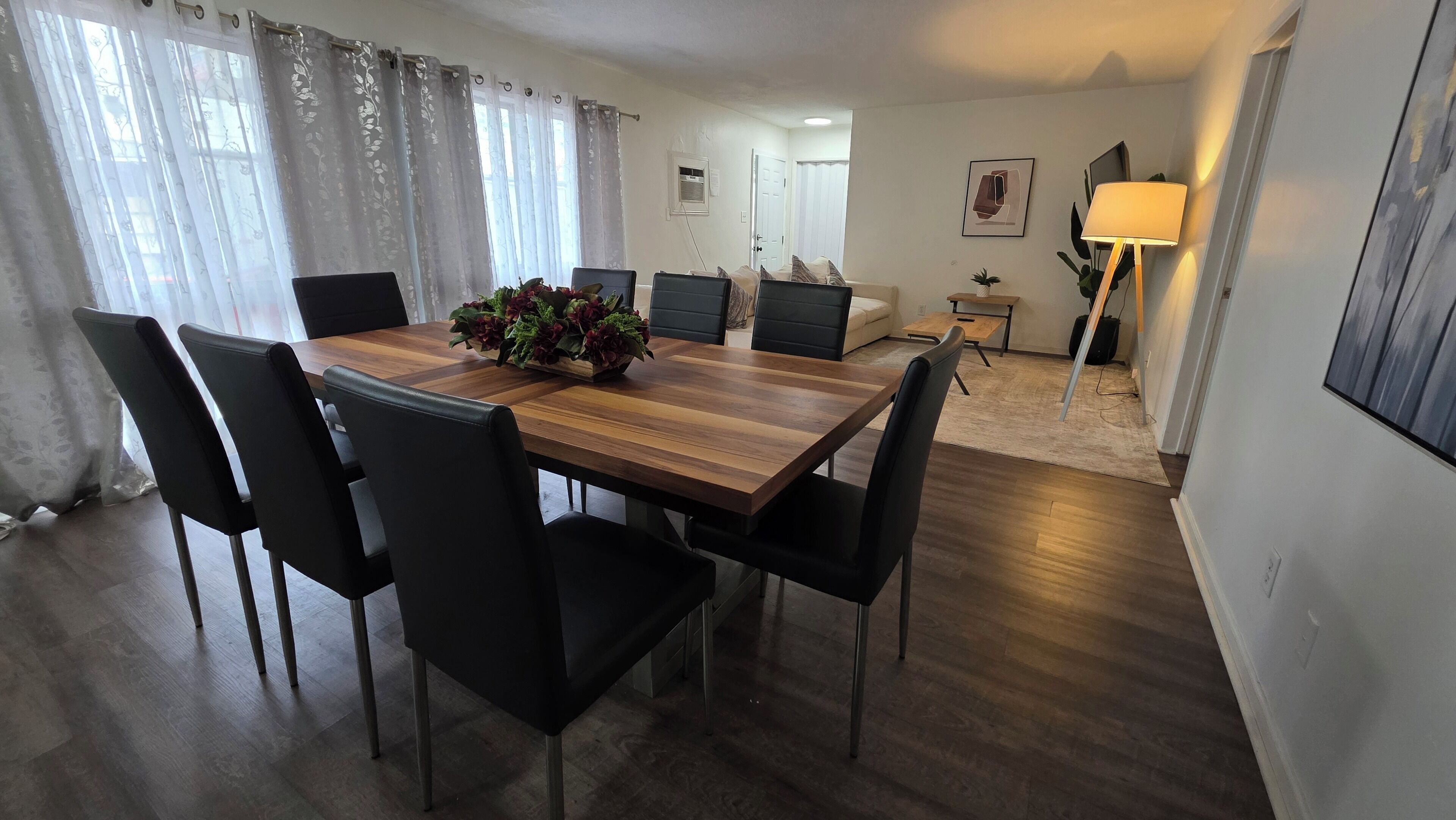 Et spiseområde med et træbord og sorte stole, en stue med en sofa og et sofabord, samt en lampe i hjørnet.