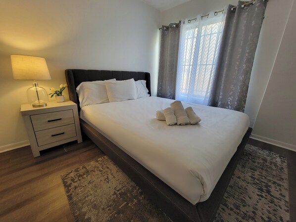 Una cama bien hecha con sábanas y toallas blancas, una lámpara de noche, una planta pequeña en maceta y una ventana con cortinas.