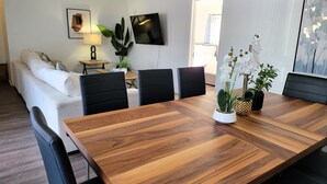 Un salon moderne avec une table à manger en bois, des chaises noires, un canapé blanc, un téléviseur fixé au mur et une plante sur la table.