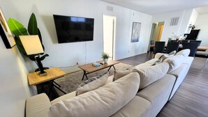 Un salon moderne comprenant un grand canapé, un téléviseur à écran plat, une table basse en bois et un coin repas avec des chaises et une table.