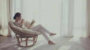 Una persona sentada en una silla de mimbre leyendo un libro en una habitación con un ventanal grande y piso alfombrado.