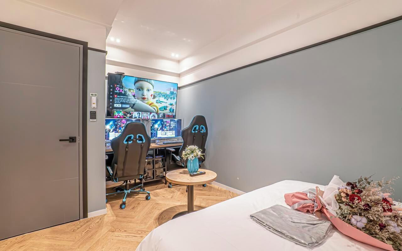 A modern hotel room with a large bed, a desk with two gaming chairs, a TV displaying a game, and a small round table with a vase.