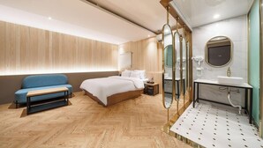 Un dormitorio moderno con suelo de madera, una cama, un sofá azul, una mesita de noche y un baño con espejo, lavabo y toallero.