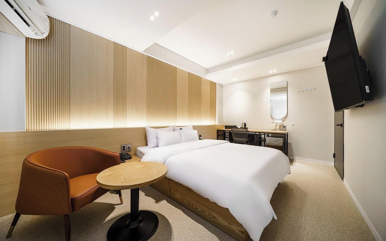 Una habitación de hotel moderna con una cama grande, una pequeña mesa redonda, un sofá, un escritorio con una silla, un televisor de pantalla plana en la pared y un muro decorativo de madera.
