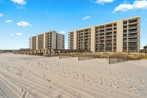 Ein Strandhotel mit mehreren Etagen, ein sandiger Strand im Vordergrund und ein klarer Himmel.