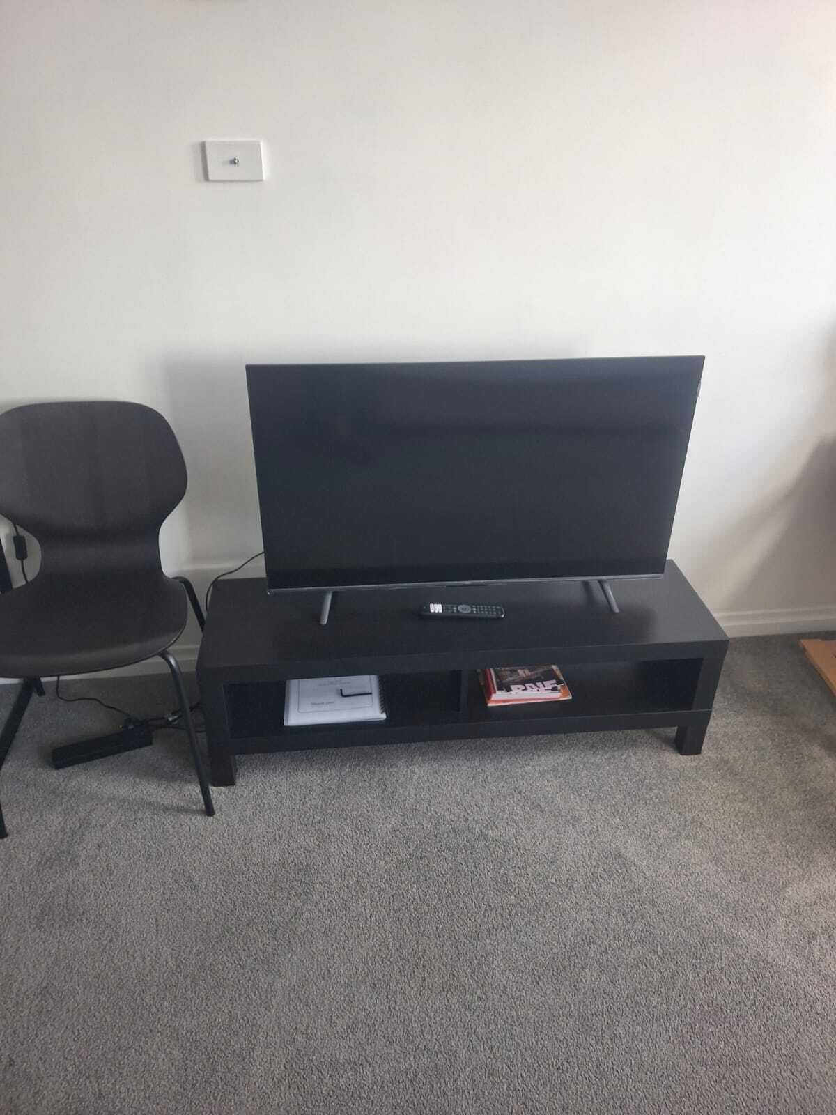 A flat-screen TV on a black stand with a remote, a black chair, and a carpeted floor.