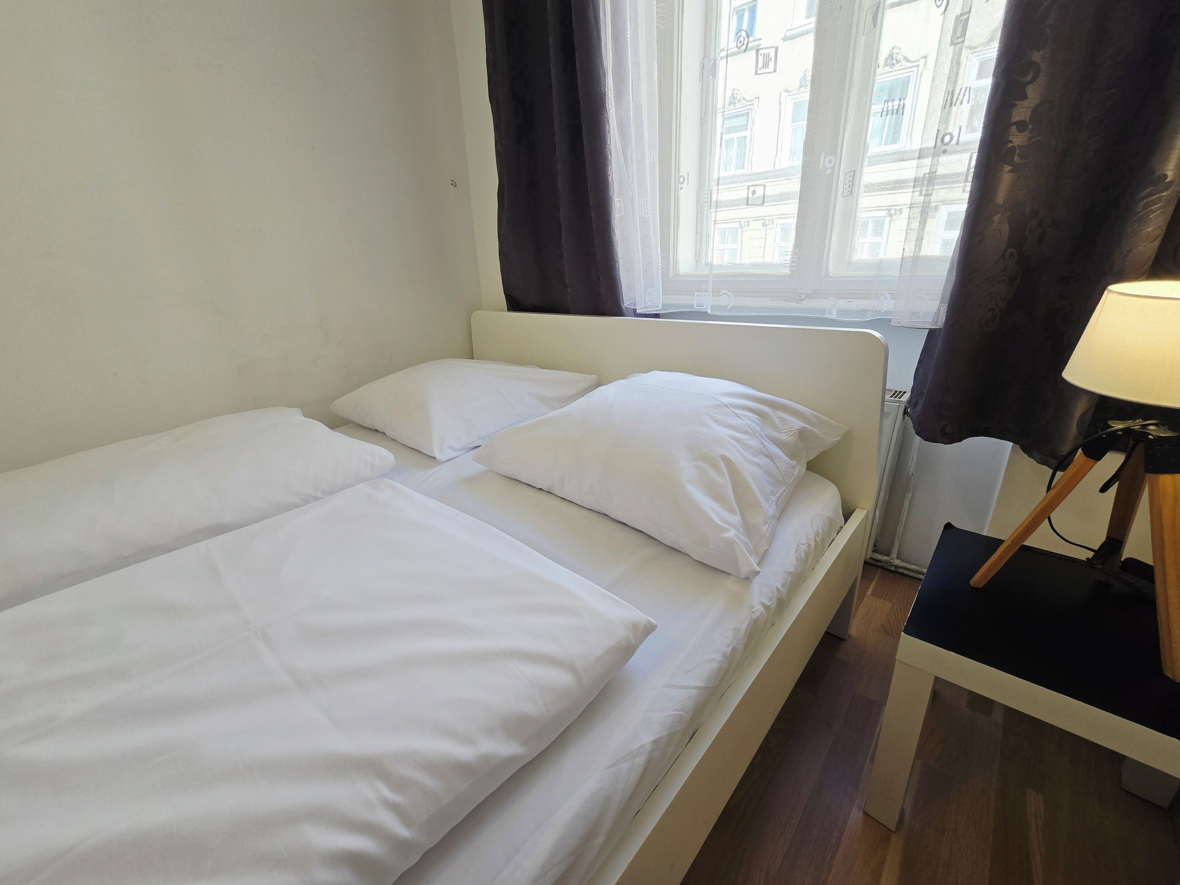 Ein Einzelbett mit weißer Bettwäsche, ein Nachttisch mit Lampe und ein Fenster mit Vorhängen.