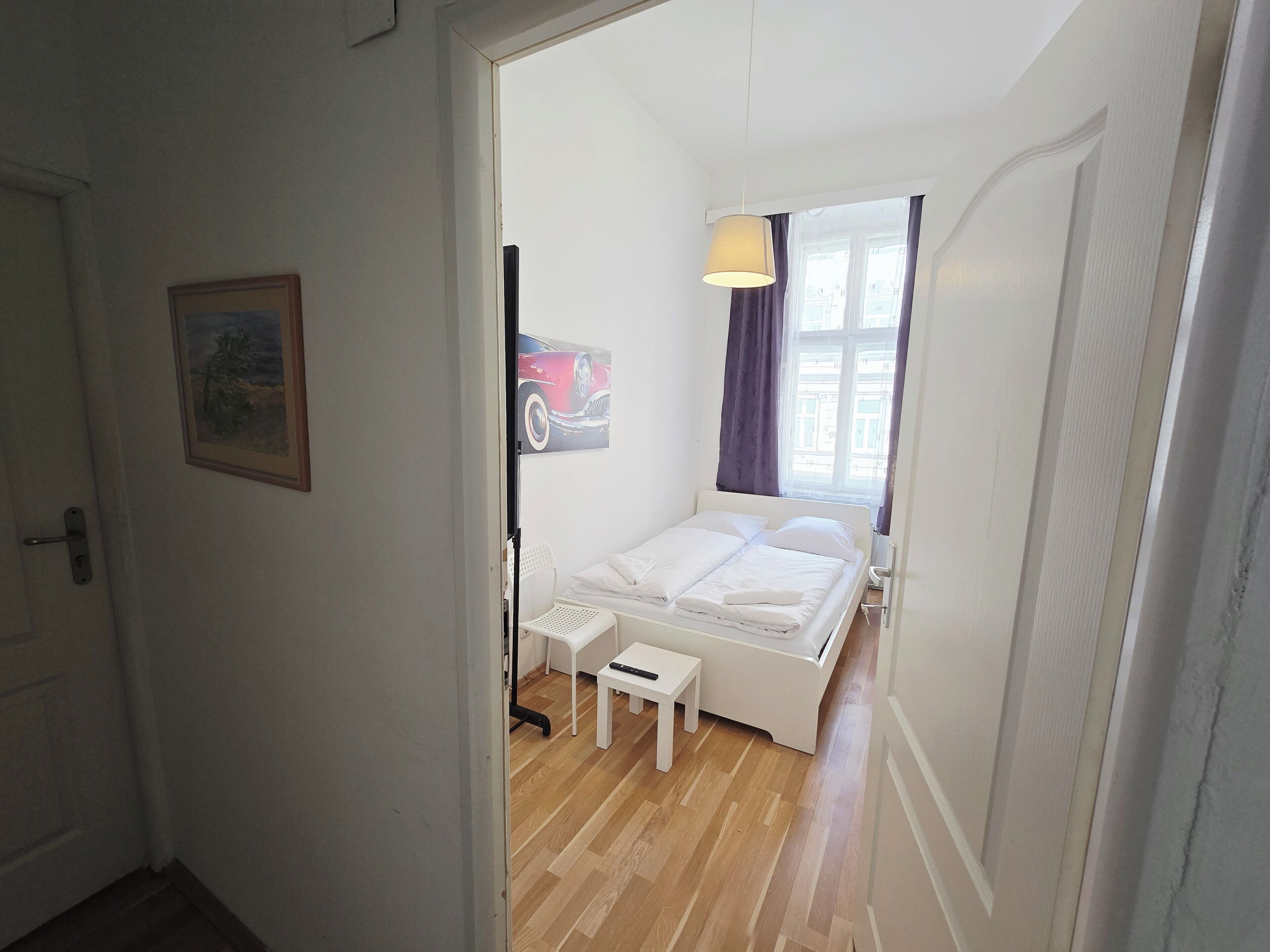 Ein Schlafzimmer mit einem Bett, einem Nachttisch, einem Sessel, einem Gemälde und einem Fenster mit Vorhängen.