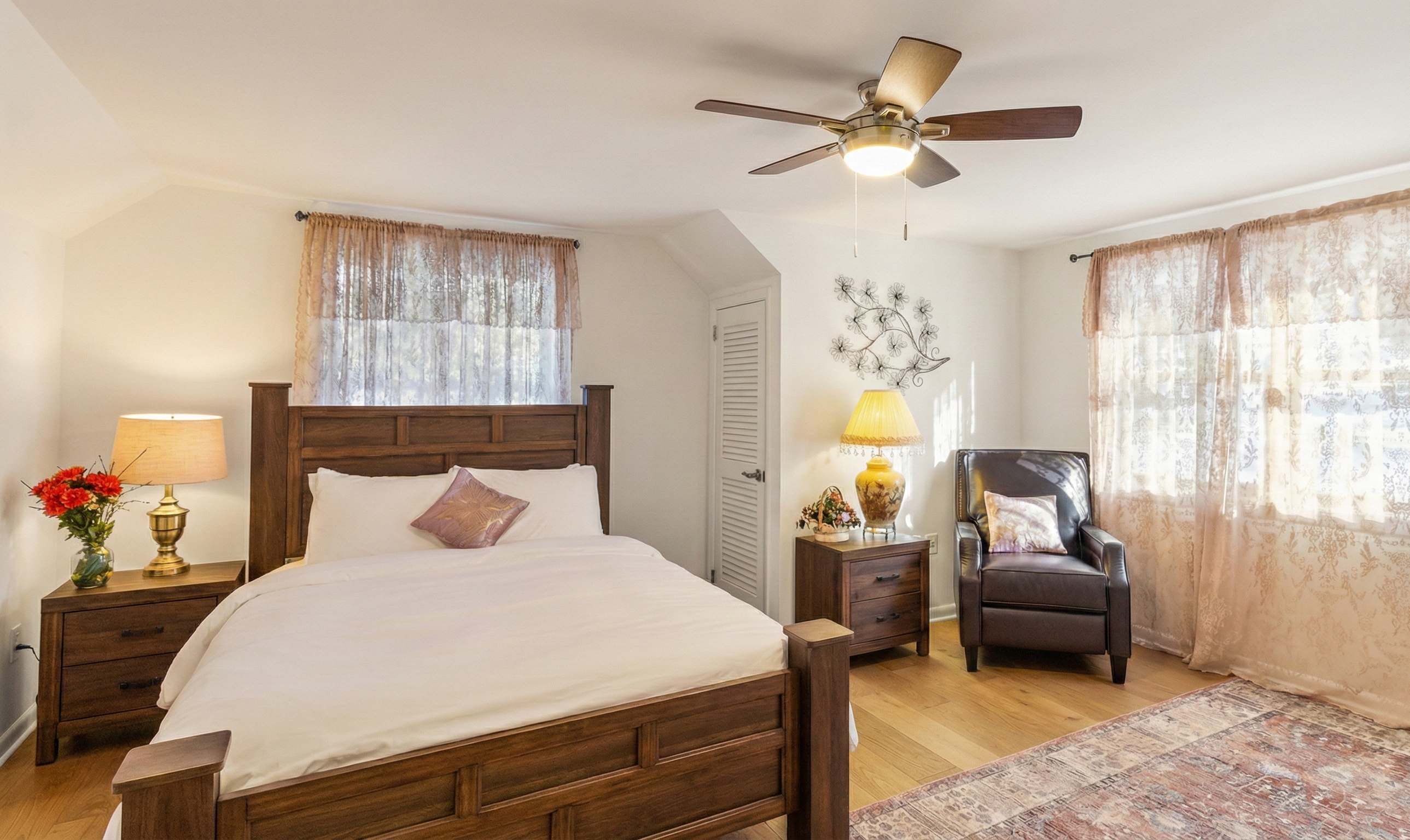 A bedroom with a wooden bed, two bedside tables with lamps, a ceiling fan, a chair, and a patterned rug.