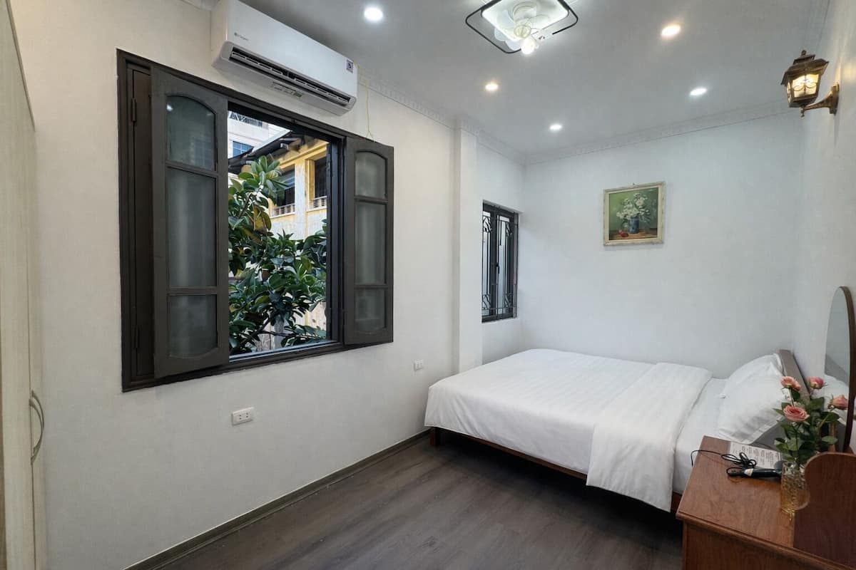 Quarto com cama, janela, ar condicionado e mesa de cabeceira com um vaso de flores.